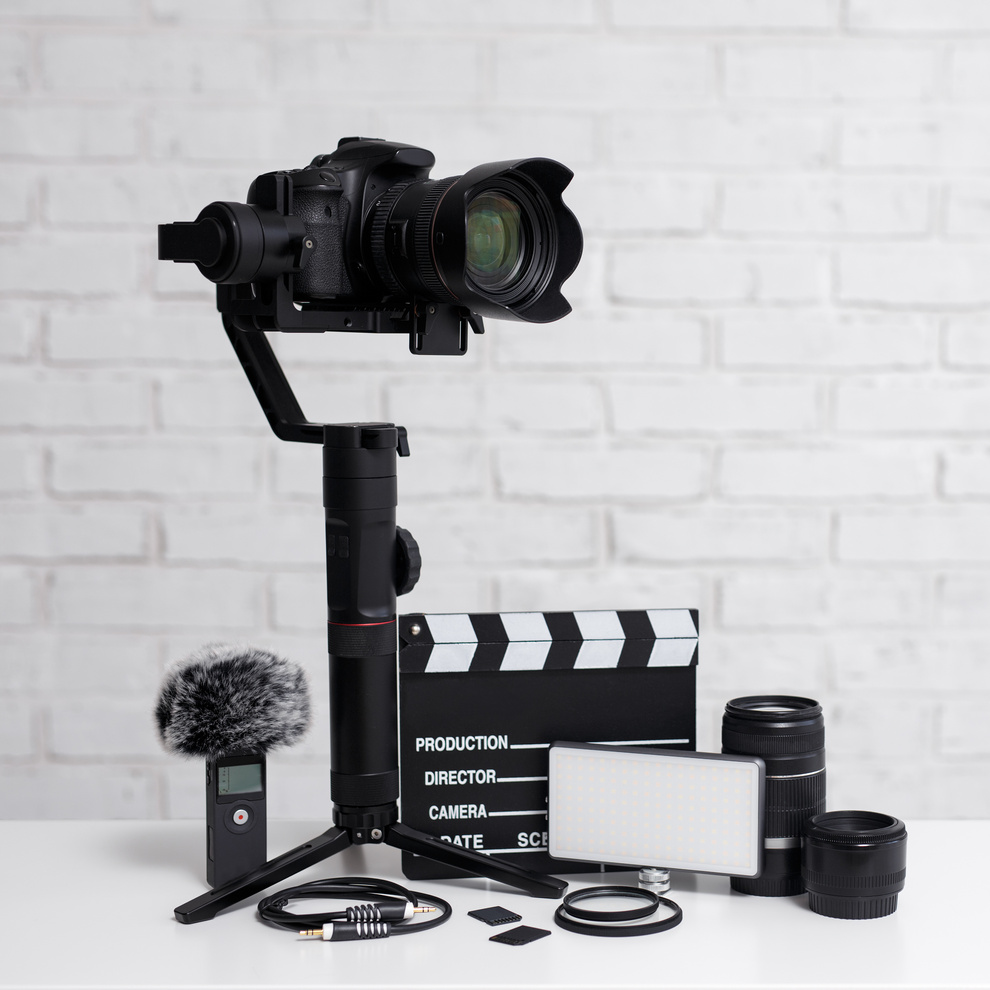 videography concept - modern dslr camera on 3-axis gimbal stabilizer, lenses, microphone, led light, clapper board and other videography equipment over white brick wall